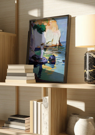 Woman by the Cliffs by Elena Avanesova in a dark frame on a wooden shelf beside books and a lamp
