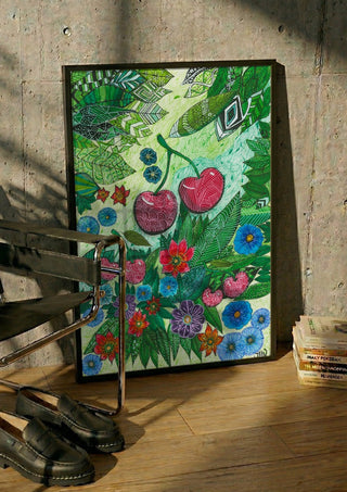 Cherries and Blue Flowers by JHN.ByHjort in a black frame leaning against a concrete wall by a steel chair