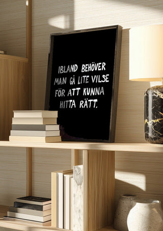 Gå vilse by Grafik bageri in a dark frame on a wooden shelf beside books and a lamp