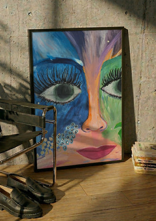 Blue and Green Face by Camilla Sabrina in a black frame leaning against a concrete wall by a steel chair