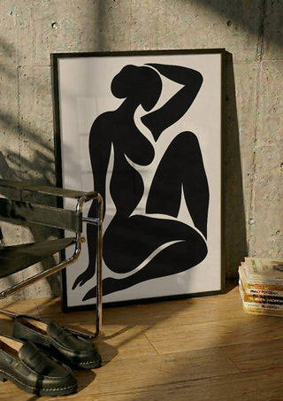 Black Nude Silhouette – Minimalist Abstract Figure Wall Art by TimlessArtworks in a black frame leaning against a concrete wall by a steel chair