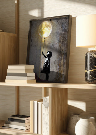 Moon Balloon by Decker Studios in a dark frame on a wooden shelf beside books and a lamp