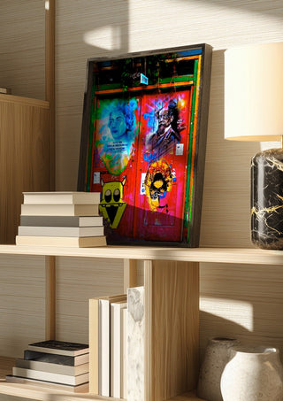 Graffiti Door by Pelecan Schneider in a dark frame on a wooden shelf beside books and a lamp