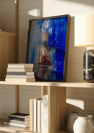 Ship in Blue by Justyna Kopania in a dark frame on a wooden shelf beside books and a lamp