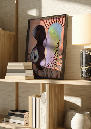 Two Heads, One Eye by Shimmering Ugly in a dark frame on a wooden shelf beside books and a lamp