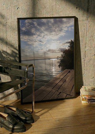 Sunset at the Lakeside Dock by Bora Kőhalmi in a black frame leaning against a concrete wall by a steel chair