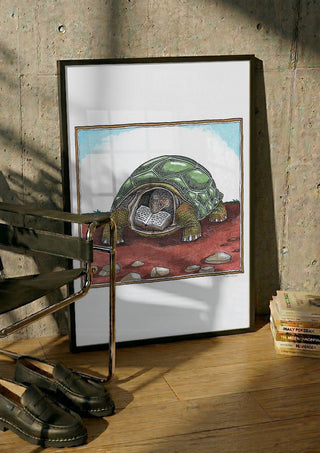 The Reading Tortoise by Waldemar Mandzel in a black frame leaning against a concrete wall by a steel chair