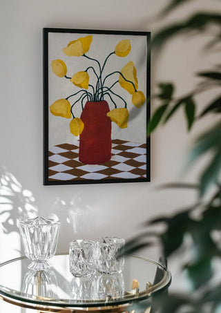 Gula blomster i terracottafärgad vas by Rebecka Hansson in a black frame above a glass table with crystal glassware