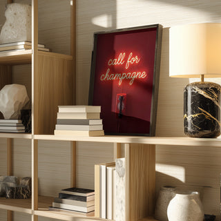 Framed neon sign reading "call for champagne" by Andreas Magnusson on a wooden shelf with books and decor.