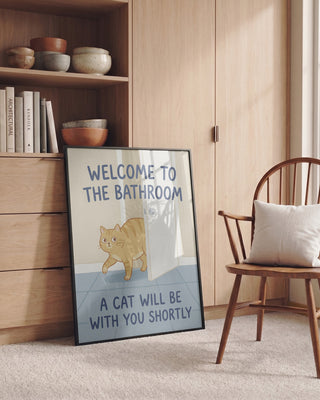 Framed Funny Bathroom Cat Welcome poster by Andreas Magnusson, leaning against a wooden cabinet in a cozy room.
