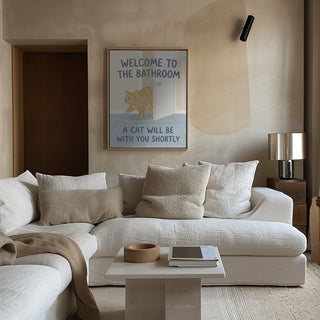 Framed "Funny Bathroom Cat Welcome" poster by Andreas Magnusson displayed above a beige sofa in a modern living room.