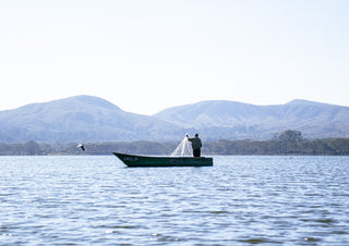 Fishermen Of Naivasha. (four) by Gloria Mwivanda - a fisherman holding his net in a green boat on a blue lake